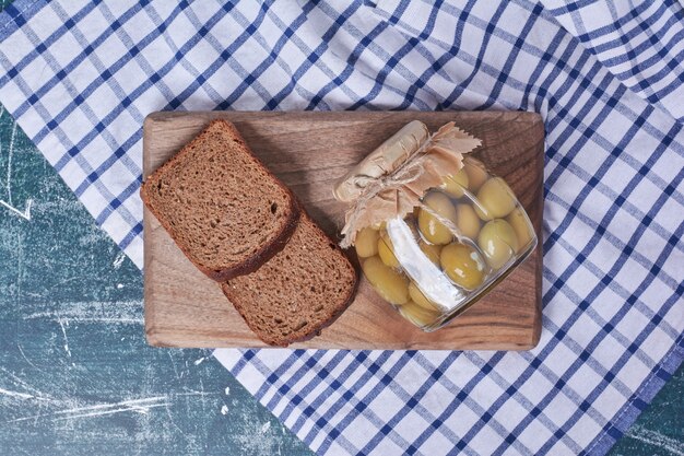 Pickled olives in glass jar with black bread slices on blue. 