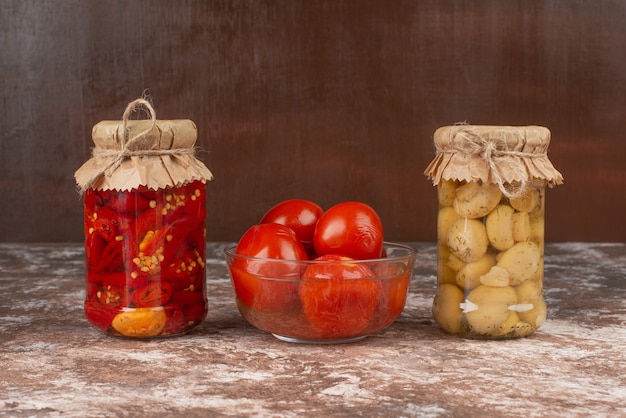 Pickled red peppers and mushrooms in a glass jar on marble table with bowl of pickled tomatoes. 