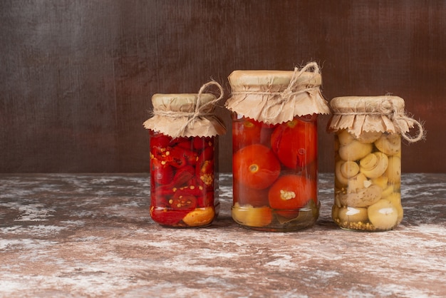 Pickled red peppers and mushrooms in a glass jar on marble table with bowl of pickled tomatoes.