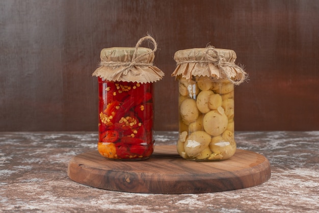 Pickled red peppers and mushrooms in a glass jar on wooden plate. 