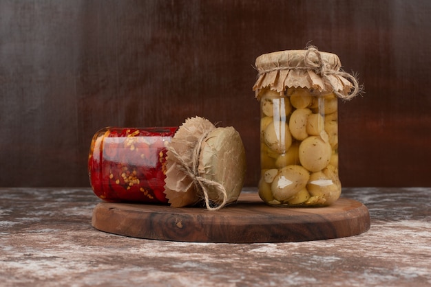 Pickled red peppers and mushrooms in a glass jar on wooden plate. 