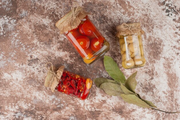 Pickled red peppers, tomatoes and mushrooms in a glass jar on marble table.