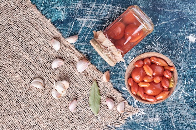 Pickled tomatoes in glass jar and bowl with garlic on blue. 