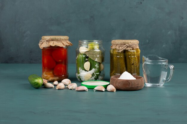 Pickled vegetables in glass jar and salt bowl on blue surface with fresh garlic. 