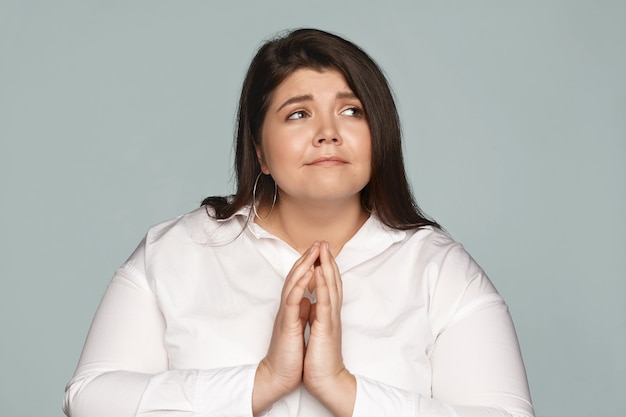 Free photo picture of adorable young chubby overweight caucasian woman in whit formal shit pressing hands together, having mournful look, begging boss to let her leave job earlier, pleading and looking up