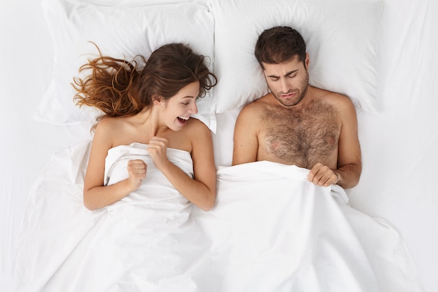 Picture of adult European bearded man and excited woman lying in bed and peeping under white blanket