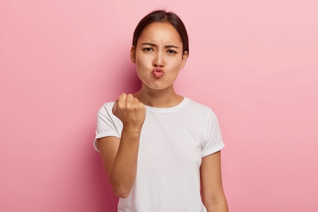 Picture of angry dissatisfied Asian woman clenches fist with displeasure, keeps lips folded, makes outraged face expression, wears white clothing, threatens you, isolated on pink wall.