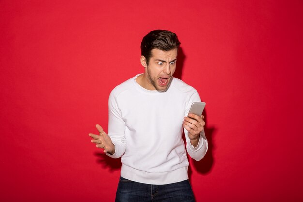 Picture of angry man in sweater screaming at smartphone over red wall