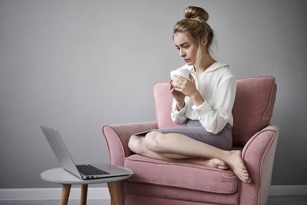 スカートと白いブラウスに身を包んだ魅力的な裸足の若い金髪の白人女性が、一般的なノートブックコンピューターで真剣にオンラインを見ながら大きなマグカップから朝のコーヒーを飲んでいる写真