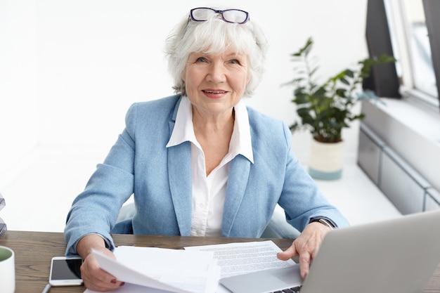Picture of attractive confident elderly mature female financial adviser with short gray hair looking with smile, studying piece of paper in her hands while working at her office desk