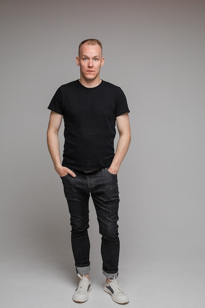 Picture of attractive man wearing a black t-shirt and jeans stands with hands in pockets isolated on grey wall