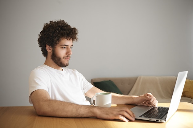 Picture of attractive stylish young man with fuzzy beard smiling watching series online or surfing internet using wifi on his generic laptop, sitting at wooden desk with mug, having coffee or tea