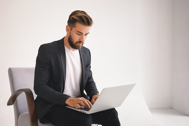 Picture of attractive successful young European bearded male entrepreneur working remotely, checking email on portable computer, having serious facial expression, focused on business issues