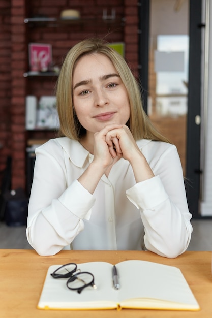 Picture of attractive successful young European female writer with blonde hair having coffee at cafe, sitting alone at wooden table with mug and open copybook, waiting for friend for lunch