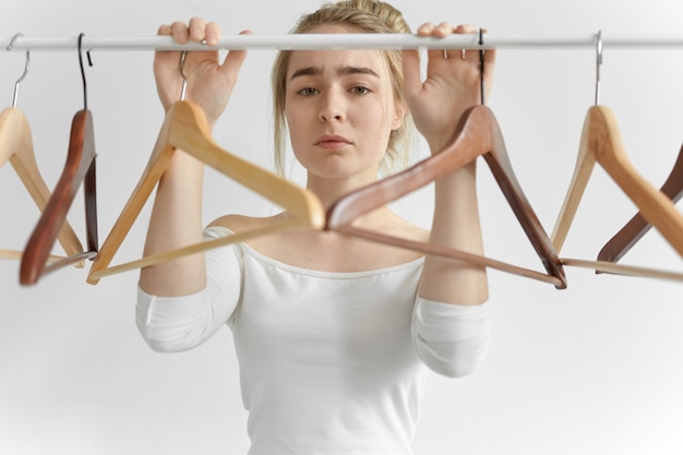 Picture of attractive young Caucasian woman in white top posing at rail with empty hangers, having sad upset facial expression because she had nothing to wear. People, clothes, style and fashion