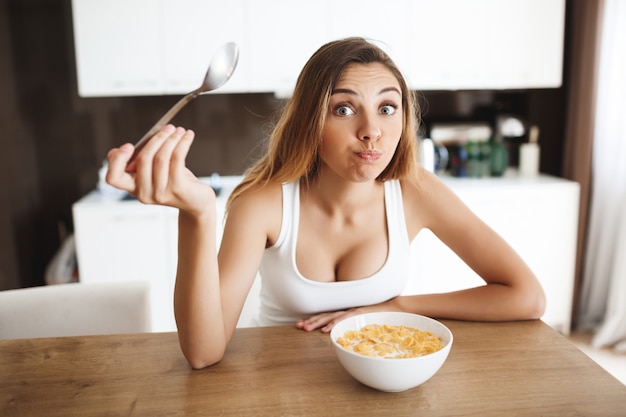 Picture of attractive young girl eating cornflakes with milk at kitchen and making fun