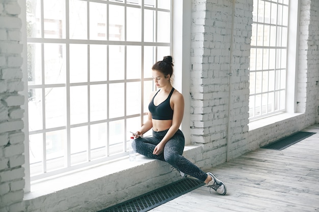 Picture of attractive young woman fitness instructor sitting on windowsill in spacious room before group workout, searching for music tracks on her mobile phone. People, sports and technology