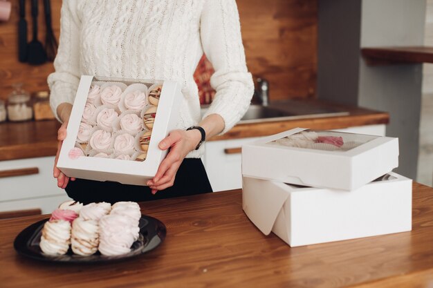 Picture of beautiful caucasian female with short dark hair in white sweater holds a white box with a lot of big white marshmallows