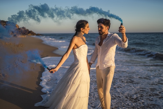 Foto gratuita foto di una bella coppia in posa con una bomba fumogena blu in spiaggia