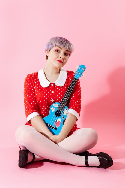 Picture of beautiful dollish girl with short light violet hair wearing red dress holding blue ukulele over pink wall