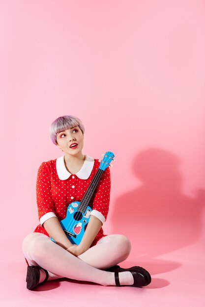 Picture of beautiful dollish girl with short light violet hair wearing red dress holding blue ukulele over pink wall