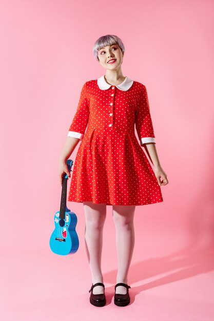 ピンクの壁に青いウクレレをかざす赤いドレスを着て短いライトバイオレット髪の美しい人形の女の子の写真