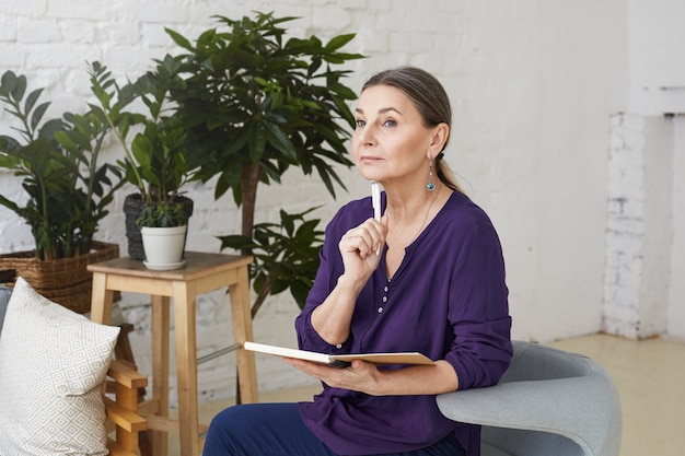 Picture of beautiful thoughtul mature 50 yeard old female writer in casual clothes sitting on comfortable chair in modern living room interior and making notes in copybook, having pensive look