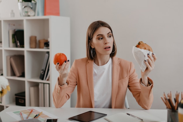 Изображение деловой женщины в розовой куртке, сидя на рабочем месте. Женщина выбирает между вкусным гамбургером и полезным яблоком.