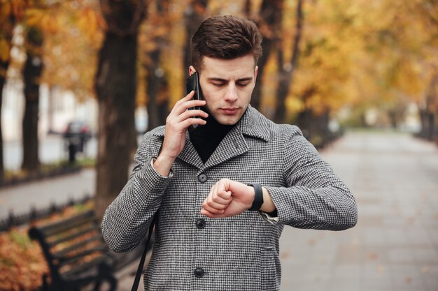 Picture of businesslike man speaking on mobile phone while going on meeting, checking time with watch on hand