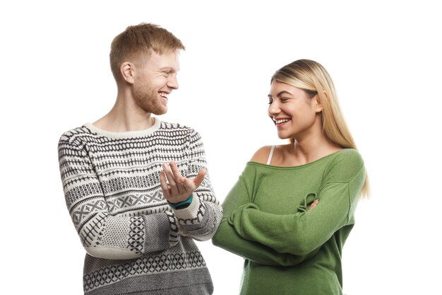 Picture of charismatic guy with stubble smiling happily while telling funny story to attractive young female with fair hair who laughing at his jokes. Cute couple talking