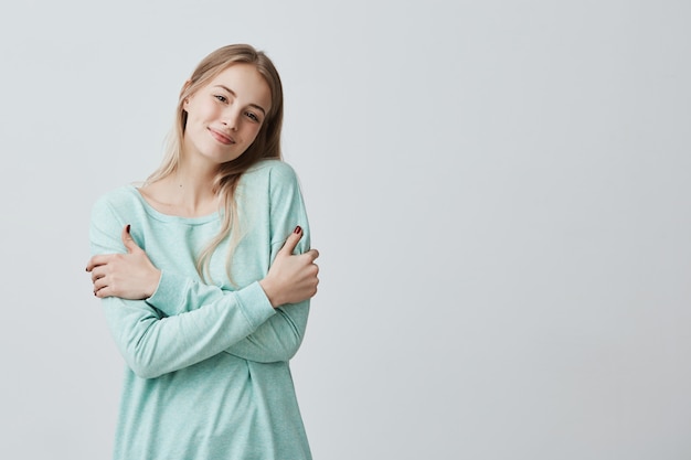 L'immagine di bella giovane femmina europea allegra si è vestita in maglione blu che sorride felicemente, abbracciandosi, avendo l'espressione allegra positiva sul suo fronte. persone, stile di vita e felicità