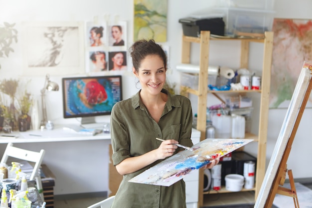 쾌활한 성공적인 젊은 여성 예술가, 팔레트 및 브러쉬 채 큰 그림 작업을 마무리 그림. 사람과 직업