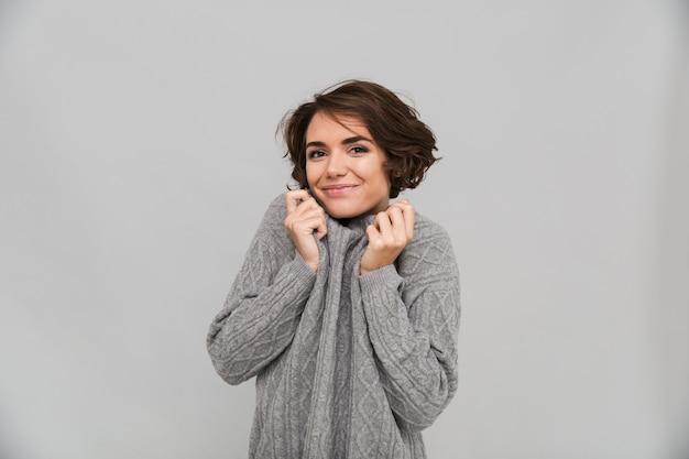 灰色の壁で分離されたセーター立っているに身を包んだ陽気な若い女性の写真。