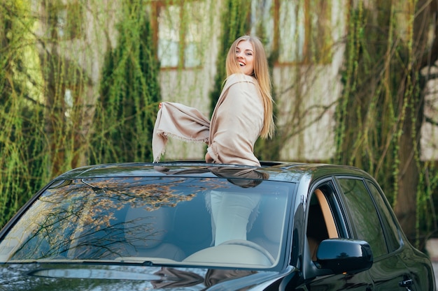 Picture of cheerful young woman wearing sunglasses and raised hands on the sunroof of the luxury car