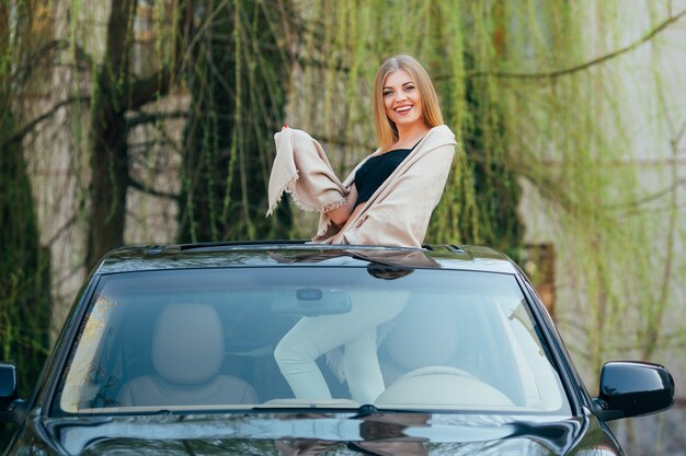 Изображение жизнерадостной молодой женщины в солнечных очках и поднятых рук на люке роскошного автомобиля