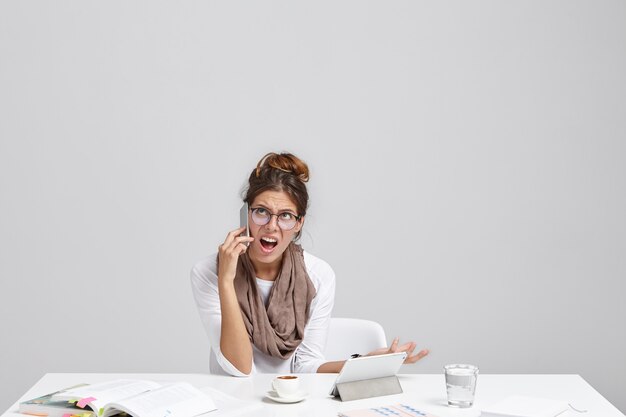 Picture of clueless emotional young Caucasian female in round glasses talking on mobile phone