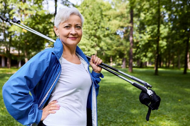 Picture of confident beautiful Caucasian female pensioner in her sixties enjoying retirement, holding trekking poles on her shoulder, going to have nordic walking outdoors, keeping hand on waist