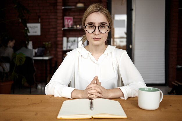 Picture of confident friendly looking young woman hr manager wearing white blouse and glasses sitting at desk with hands clasped during job interview, asking questions and listening attentively