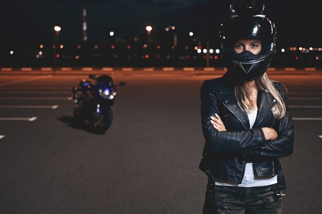 Picture of confident self determined young female rider in safety helmet standing in parking lot keeping arms crossed and looking, going to have ride on motorcycle around night city