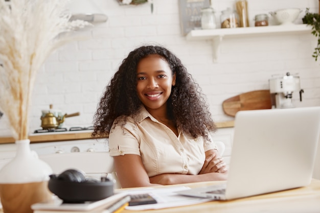 Picture of cute stylish young African American woman accountant with confident toothy smile working remotely on laptop computer, doing finances in kitchen. Technology, occupation and freelance