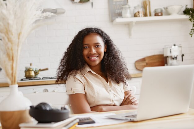 キッチンで財政をしているラップトップコンピューターでリモートで働いている自信を持って歯を見せる笑顔でかわいいスタイリッシュな若いアフリカ系アメリカ人の女性会計士の写真。テクノロジー、職業、フリーランス