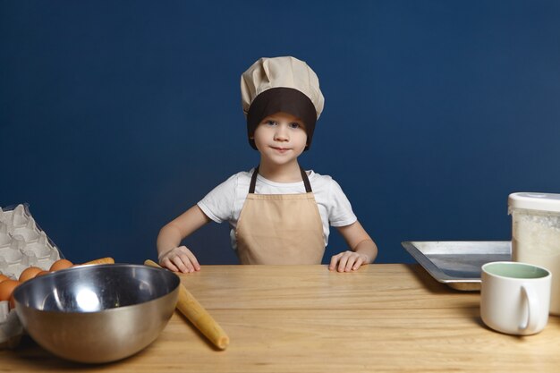 Изображение решительного взволнованного маленького мальчика в униформе шеф-повара, стоящего за кухонным столом с металлической миской