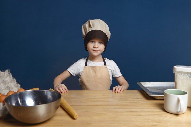 Picture of determined excited little boy wearing chef uniform standing at kitchen table with metal bowl