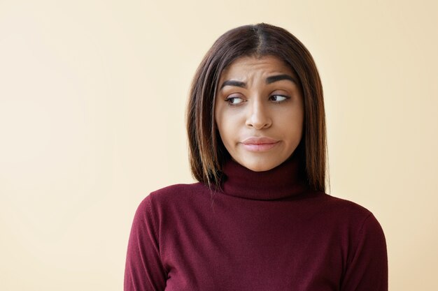 Picture of disappointed young dark skinned brunette female looking sideways with eyes expressing sorrow and disappointment, feeling upset while having stress at work or problems in personal life