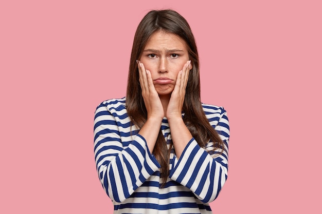 Picture of dissatisfied Caucasian teenager purses lips, keeps both hands on cheeks