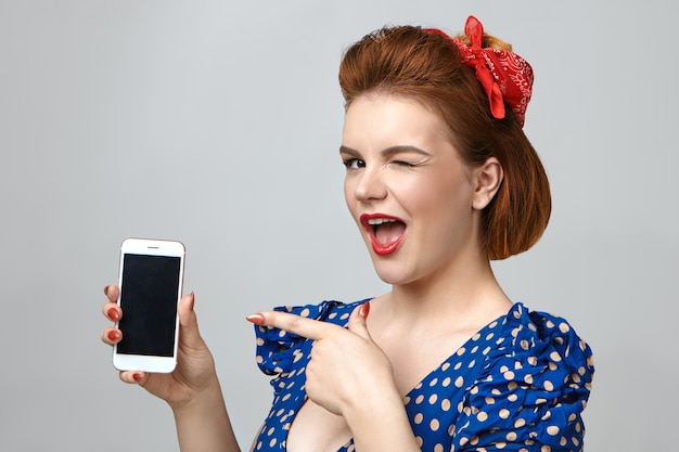 Picture of elegant emotional young woman dressed like pin up girl winking playfully at camera and pointing index finger