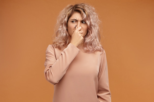 Picture of emotional displeased young European woman pinching her nose because of bad smell or gross stink. Stylish teenage girl can't stand dirty socks odour