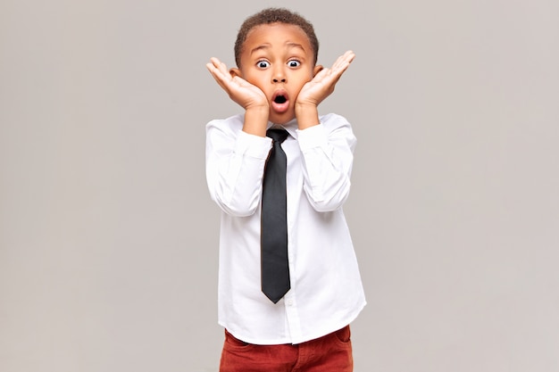 Picture of emotional funny surprised African American schoolboy in shirt and tie holding hands at his face, widening eyes and opening mouth widely, being shocked with astonishing unexpected news