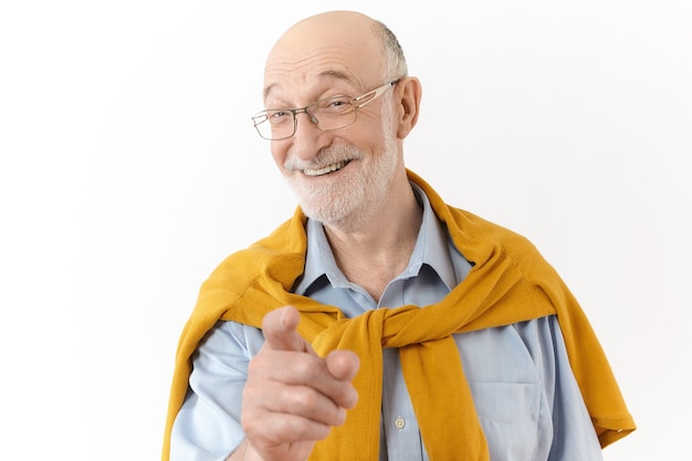 Картина эмоционального красивого старшего мужчины с лысой головой и серой щетиной, широко улыбающегося и указывающего указательным пальцем на камеру, смеющегося над забавной историей или шуткой, позирующего изолированно на белой стене студии