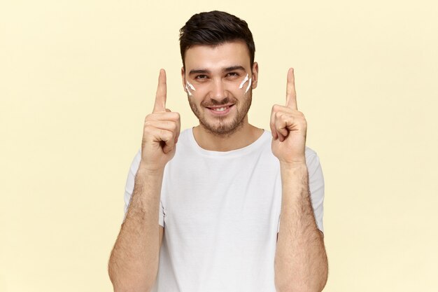 Picture of emotional overjoyed young man with moisturizer on cheeks smiling at camera, pointing fore fingers up. Cute guy raising finger, having great idea